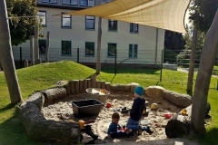 Der große Sandkasten ist umrandet von Baumstämmen, Findlingen und Hölzern. Im Hintergrund der Rutschhügel für erste Rutschpartien im Winter.